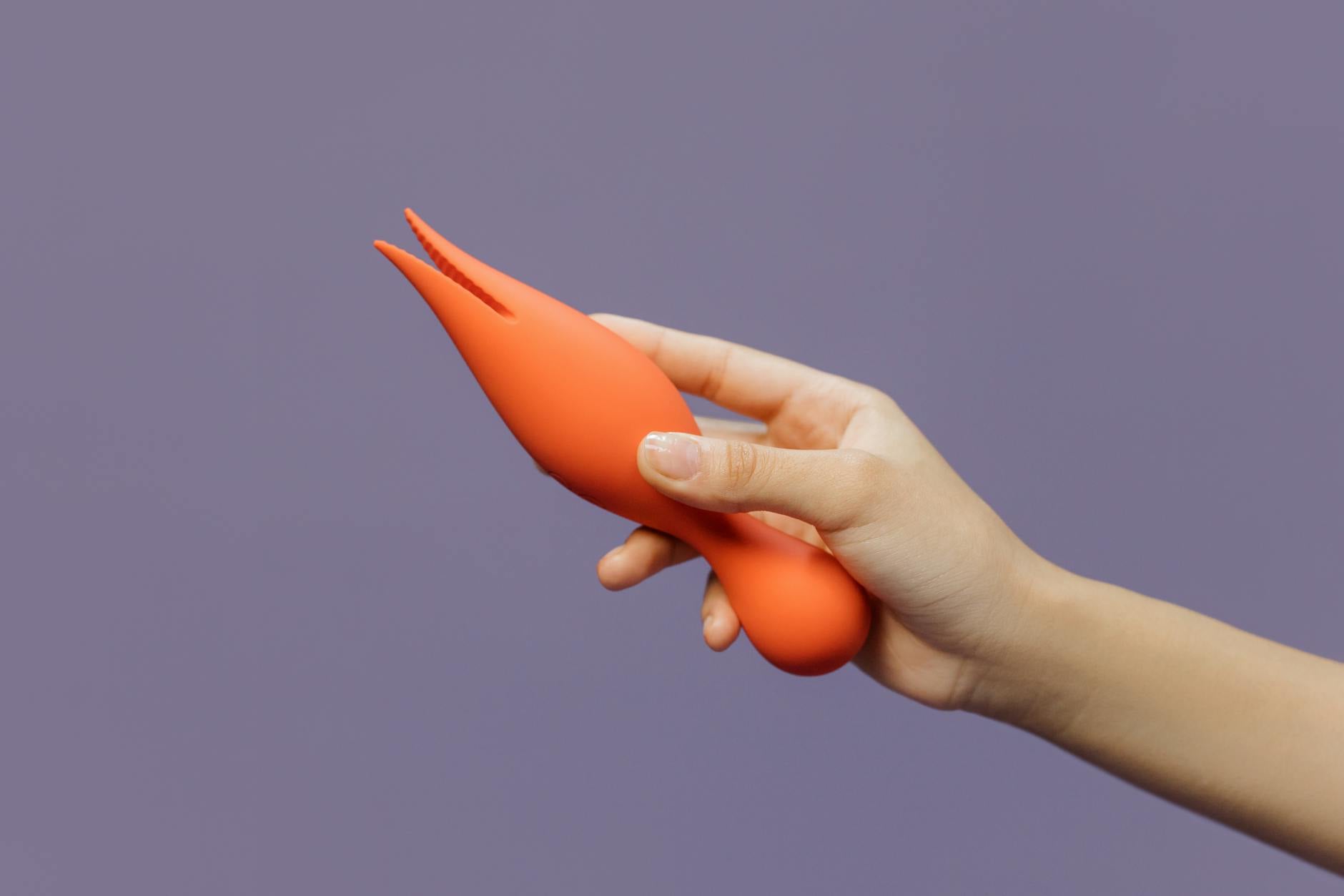 A hand holding an orange clitoral vibrator against a minimalistic purple backdrop