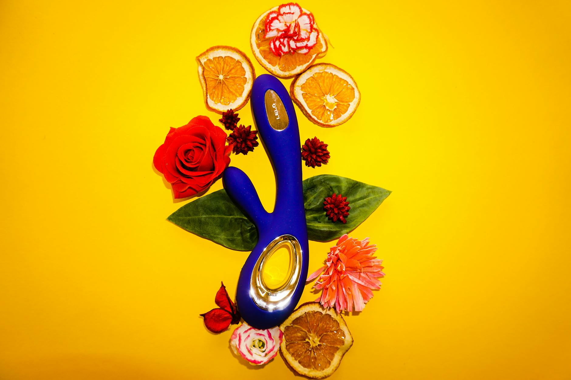 A blue clitoral vibrator surrounded by fresh citrus and flowers on a yellow background, representing extended pleasure with Lemon vibrators